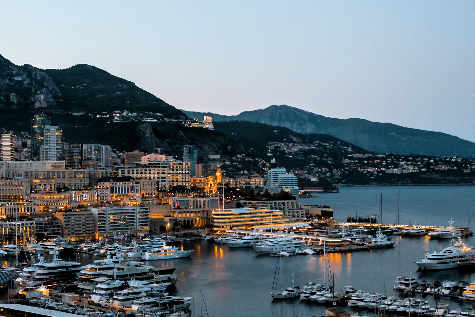 Monaco's harbour at twilight from the hillside, superyachts lit along the quays below the mountains