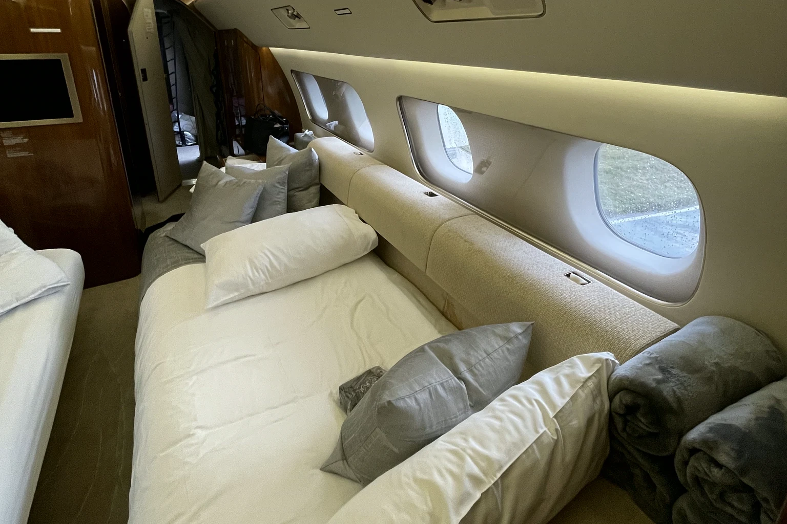 A divan along the cabin wall of a private jet made up as a bed with white sheets, pillows and rolled grey blankets, oval windows above