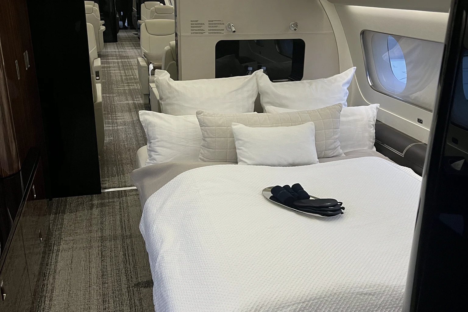 A made-up double bed with white pillows and a tray of slippers in a private jet cabin, dark wood cabinets on the left and seats beyond