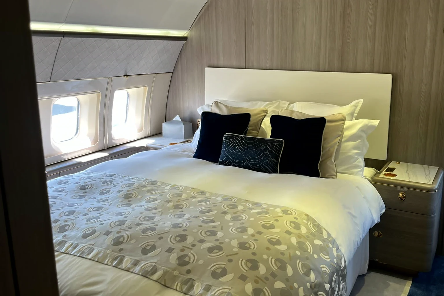 A double bed with a white headboard, navy cushions and a patterned throw in a private jet bedroom, two windows on the left