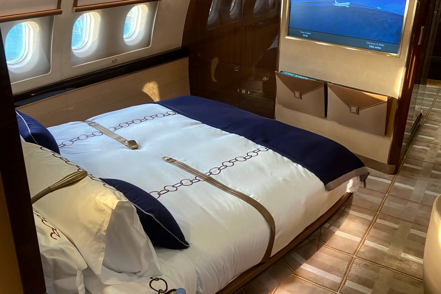 A double bed with a white and navy cover in a private jet bedroom, wood panelling, three windows and a moving-map screen