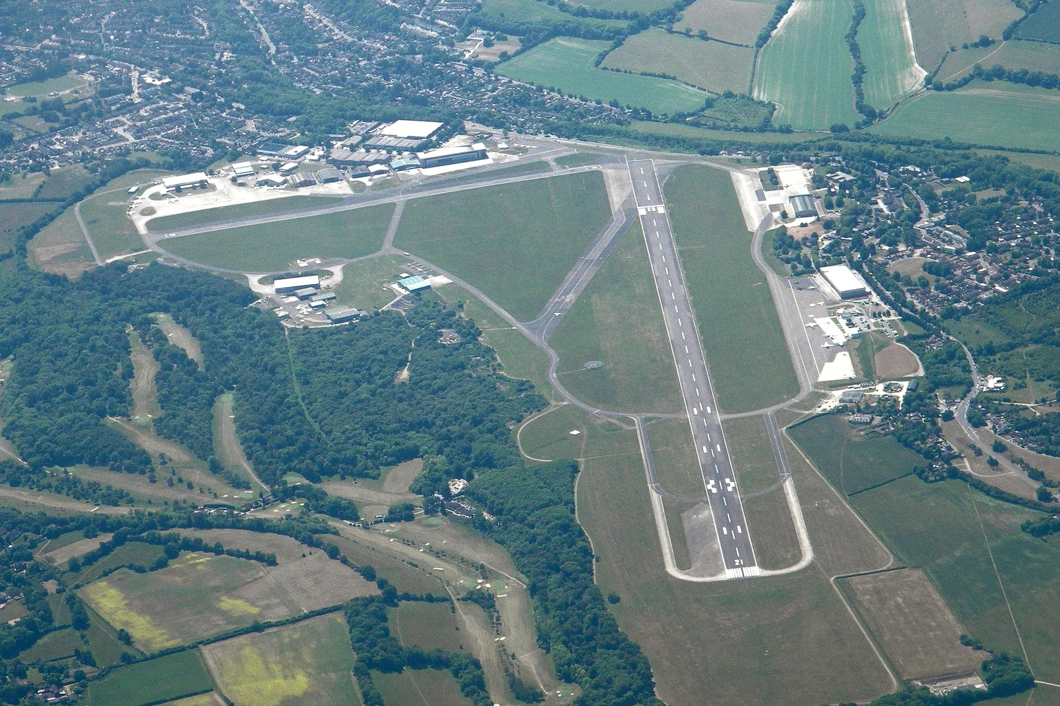 Biggin Hill Airport from the air: one runway, the hangars and terminals beside it, fields and woods around