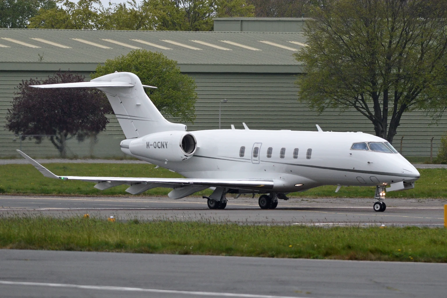 A white Bombardier Challenger 350 taxiing in front of a green hangar at Biggin Hill