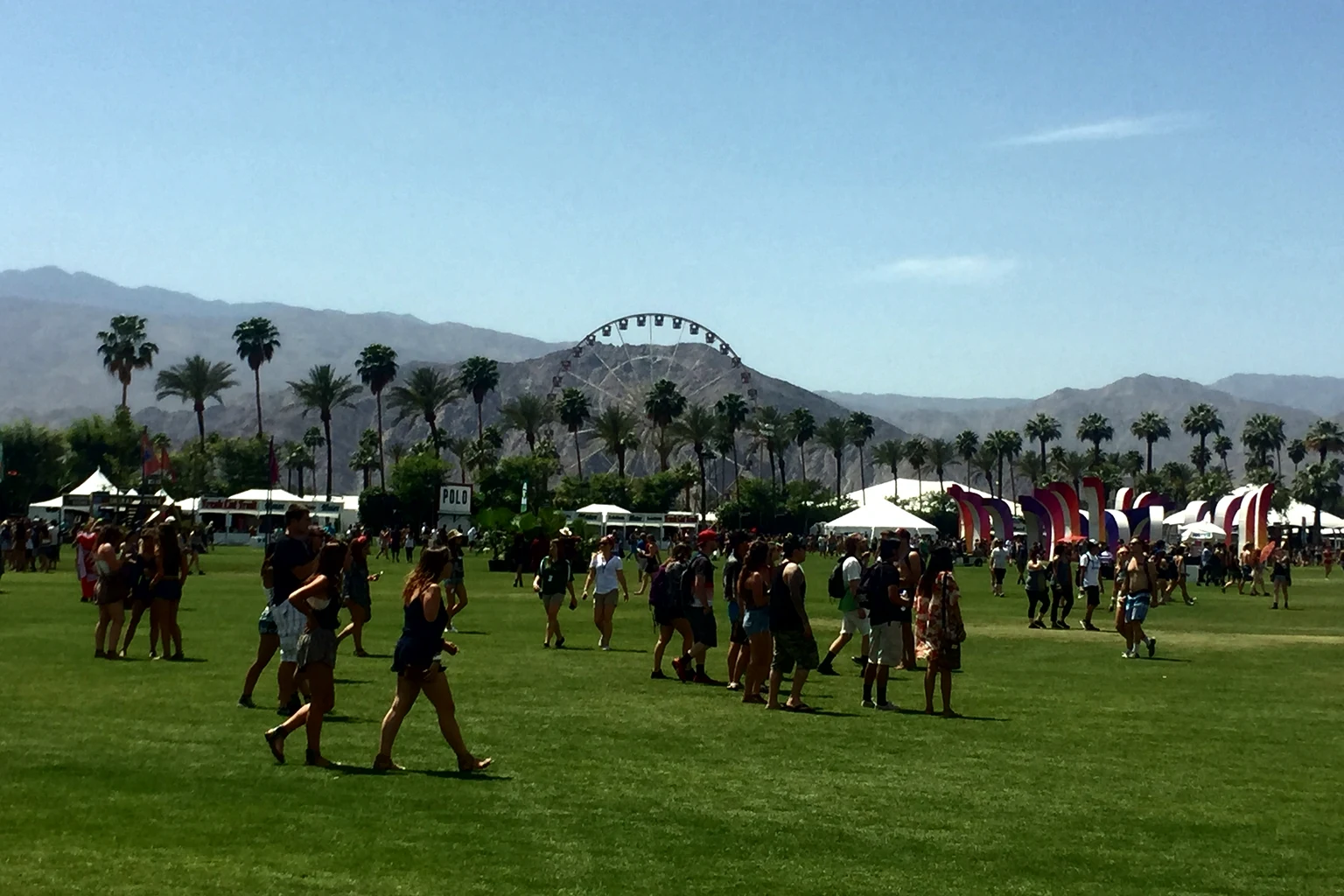 Festival-goers crossing the lawn of the Empire Polo Club in Indio, the Coachella Ferris wheel and the palms in front of the mountains