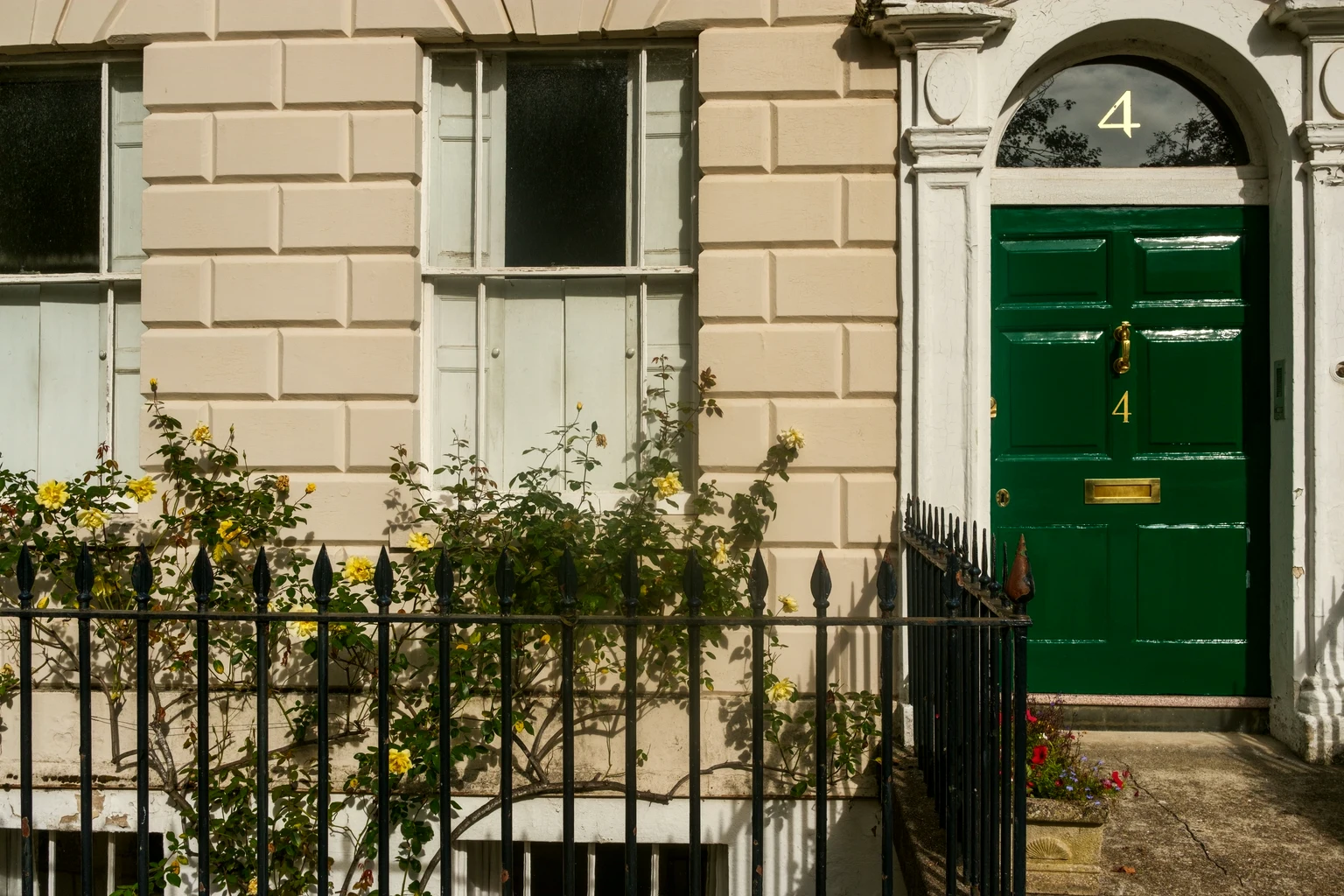 A glossy green Georgian front door with brass fittings, behind black railings and yellow roses