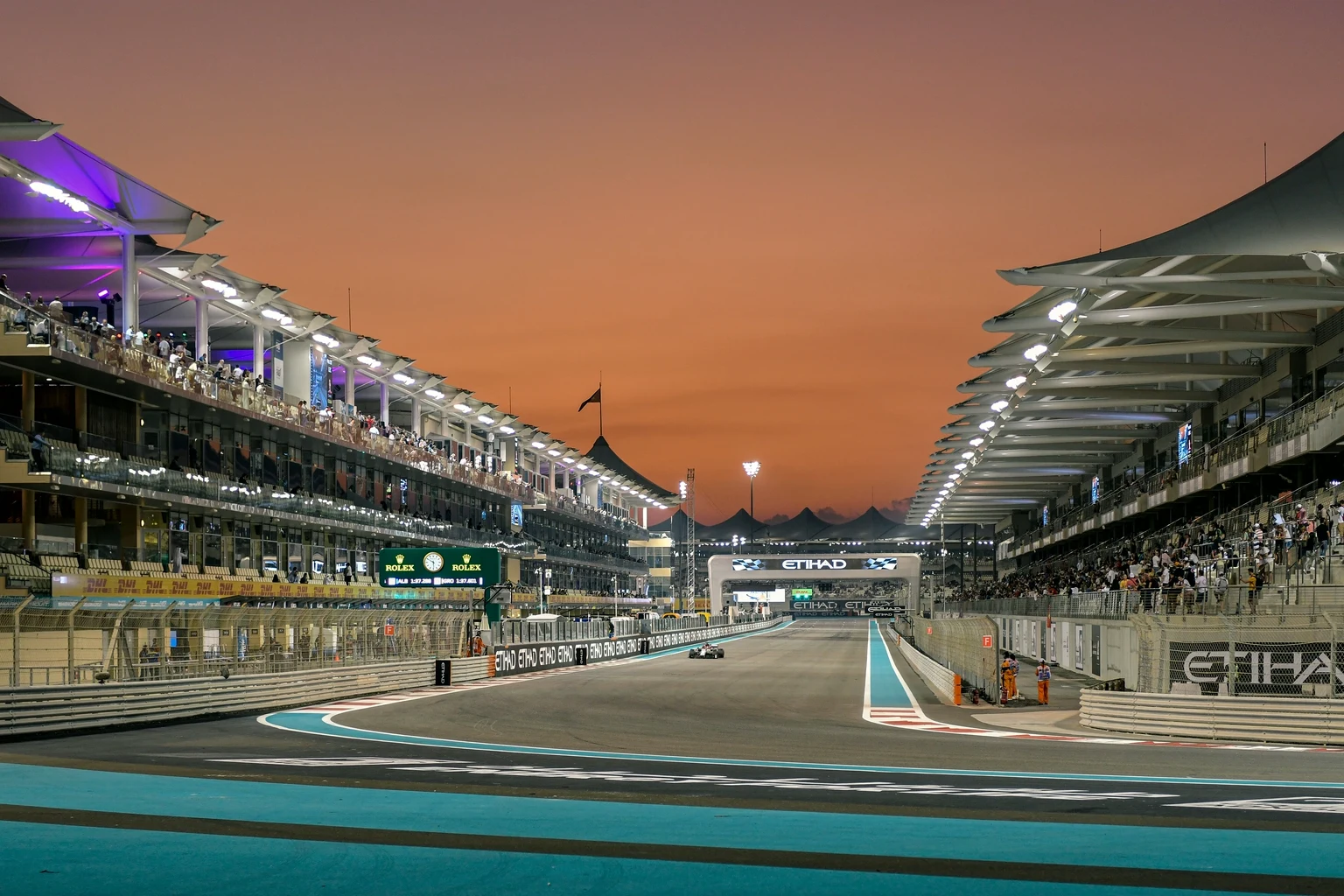 The start-finish straight at Yas Marina Circuit at sunset, the grandstands lit on both sides and a single car in the distance