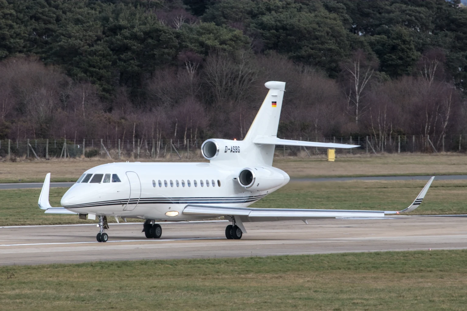 A white Dassault Falcon 900EX taxiing at Farnborough in front of pine woods