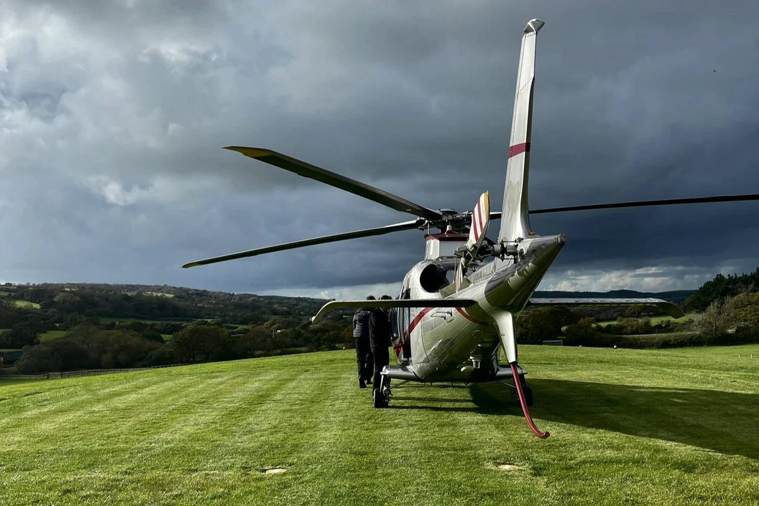 A silver AgustaWestland AW109 landed on a country-house lawn under a storm sky, its passengers walking up to the door