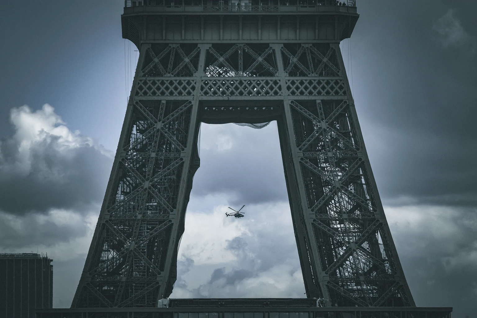 A helicopter framed between the legs of the Eiffel Tower under a stormy sky