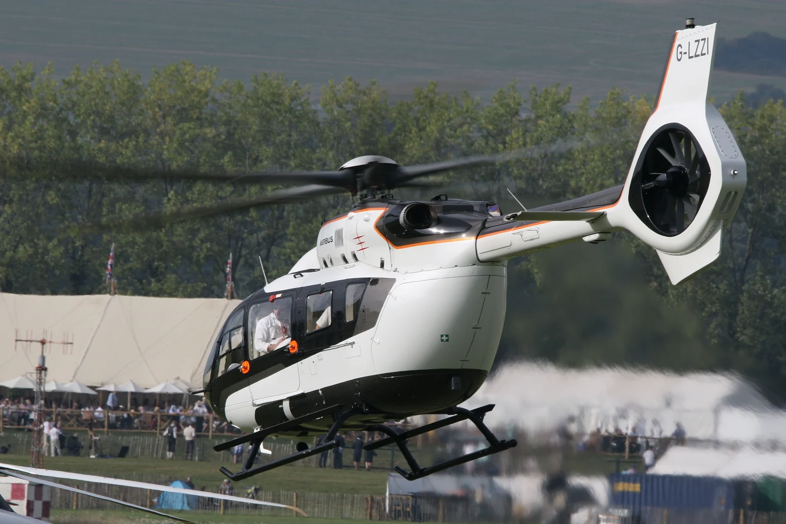 A white Airbus H145 helicopter coming in to land past the hospitality tents at the Goodwood Revival