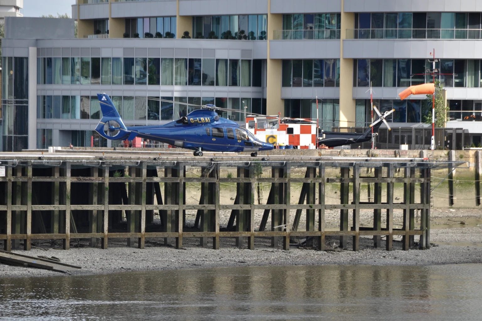 A blue Eurocopter Dauphin on the landing platform of The London Heliport, raised on timber piles above the Thames at low tide