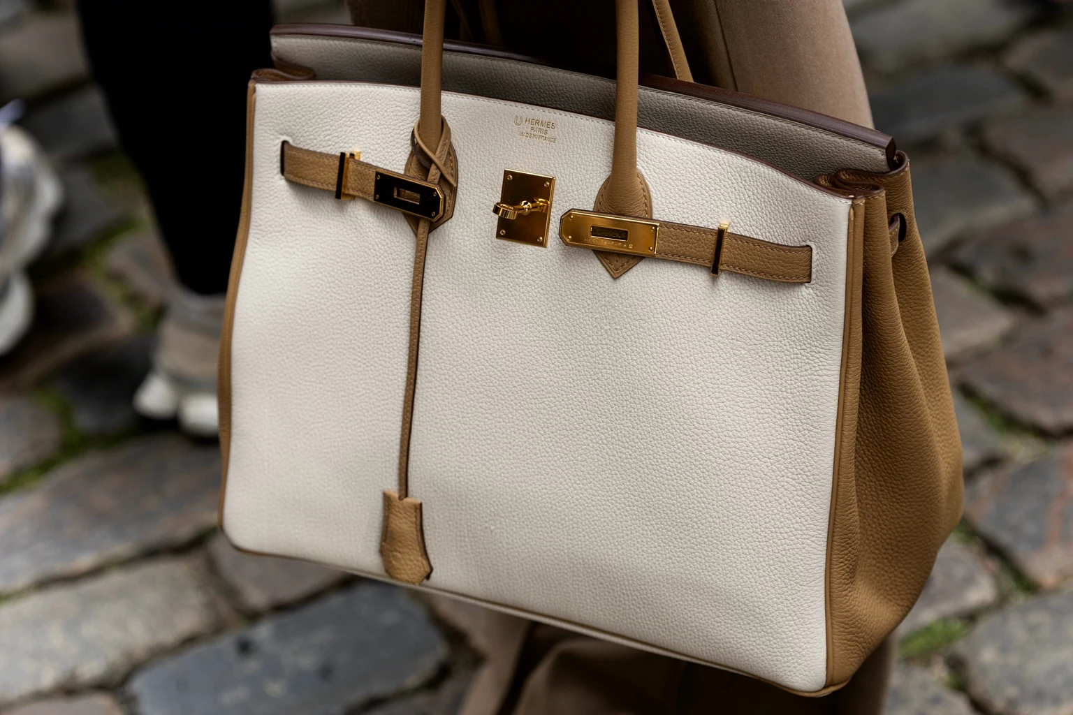 A two-tone Hermès Birkin in white and tan leather with gold hardware, carried on a cobbled street