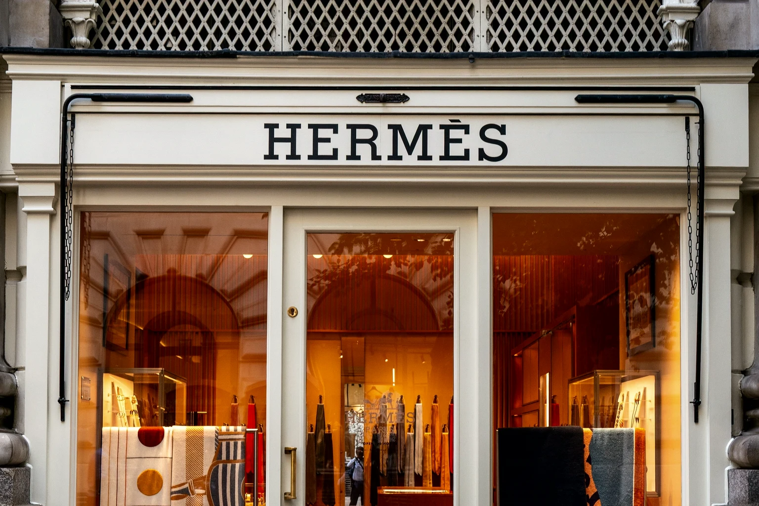 The front of an Hermès boutique, its name over the door and silk and cashmere pieces lit in the windows