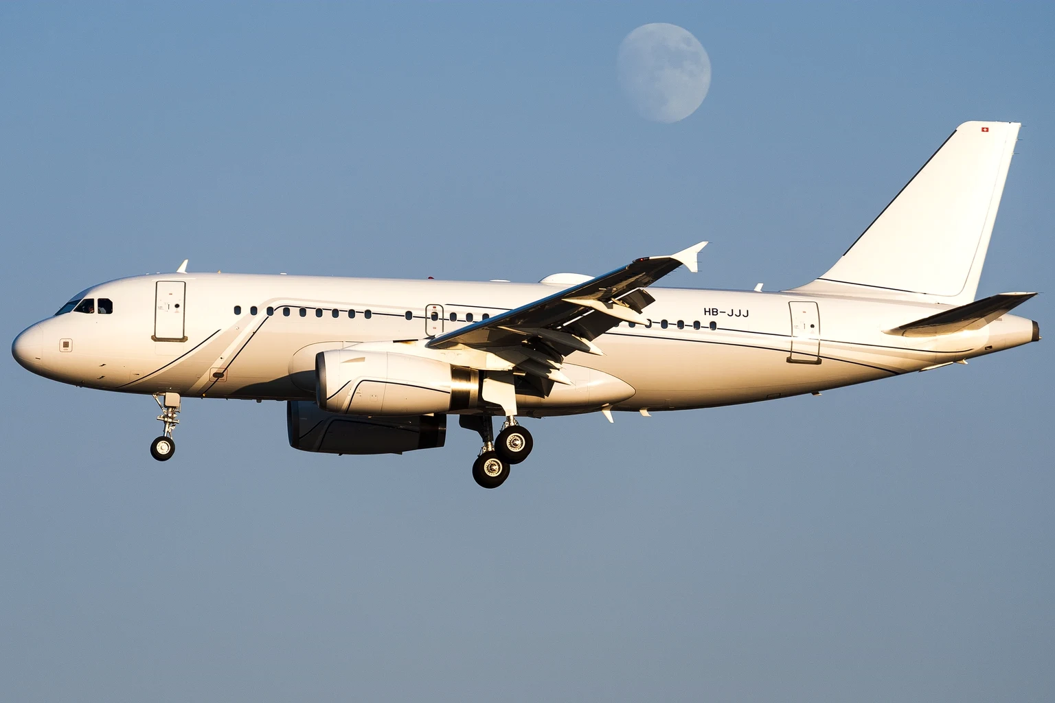A plain white Airbus ACJ319 landing in warm late light with the moon in the sky.
