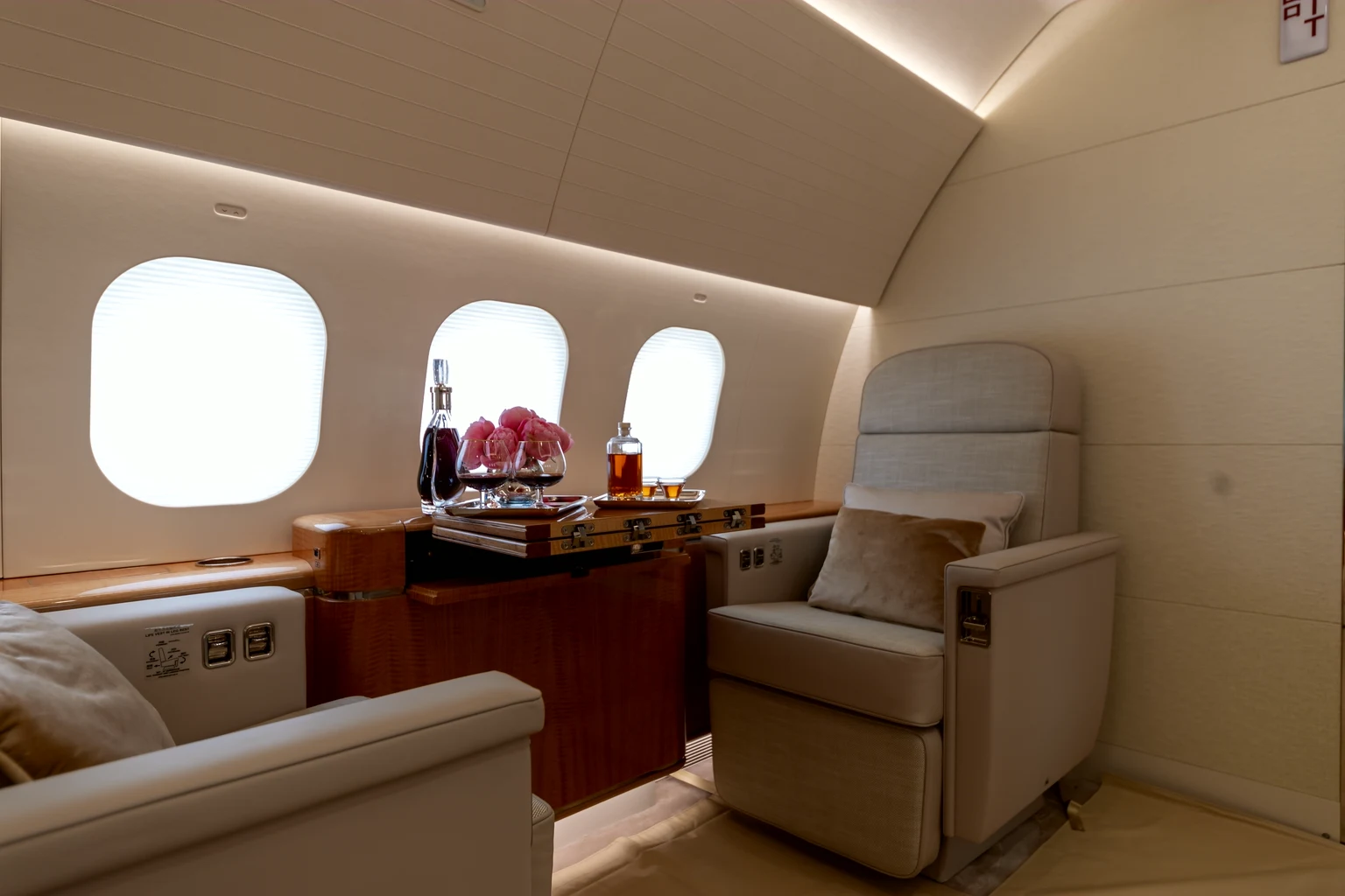 A cream club chair beside a walnut credenza set with pink roses and a decanter under warm cove light in an ACJ319neo.