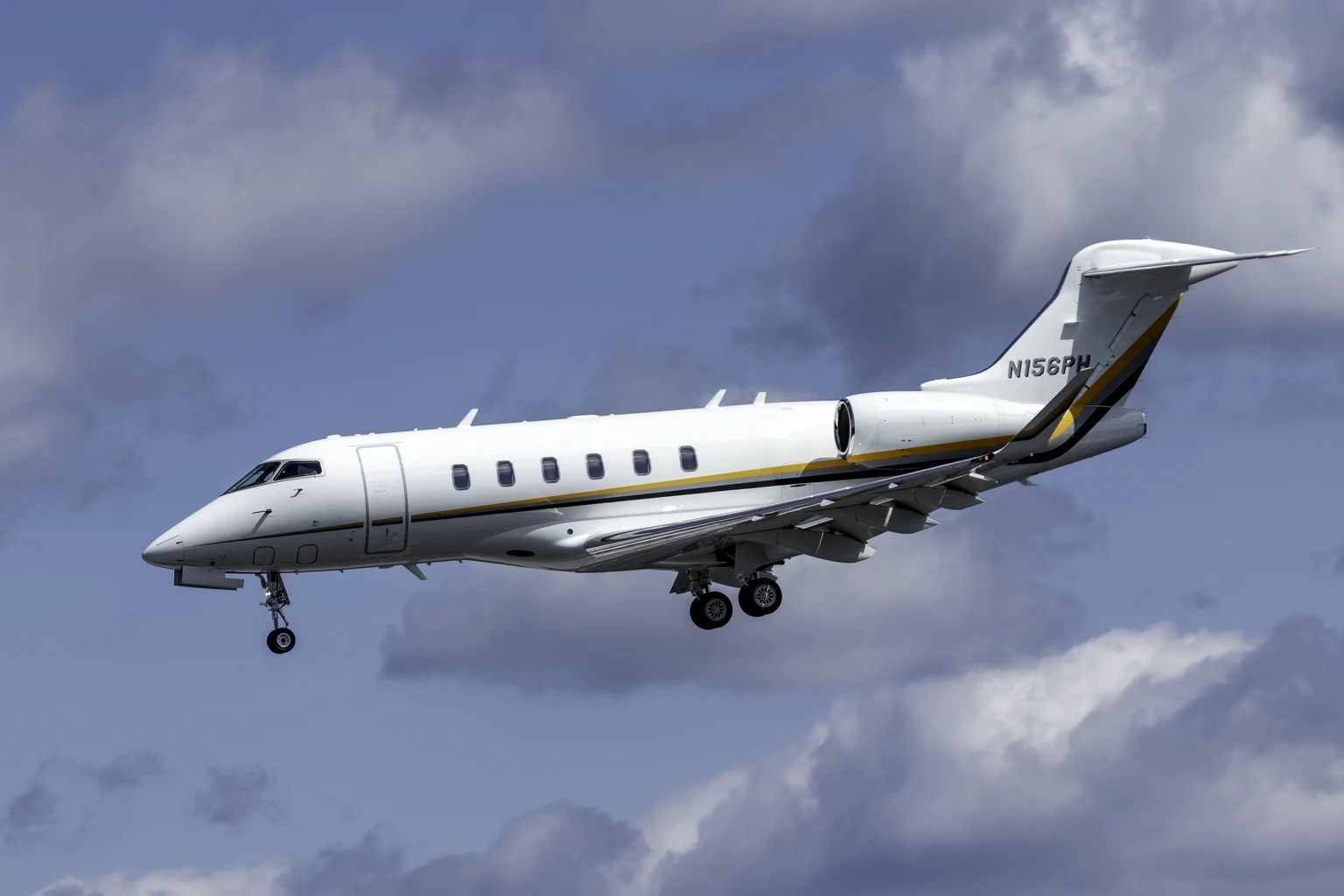 A white Bombardier Challenger 350 with a gold and black cheatline on approach under a broken-cloud blue sky, three-quarter from below.