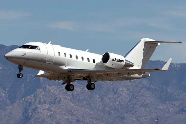 A white Bombardier Challenger 605 on approach, three-quarter from the front, with brown desert mountains and a blue sky behind.