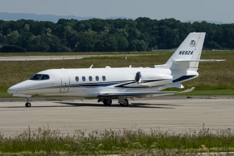 A white Cessna Citation Latitude with blue swooshes and the Cessna logo on its tail, taxiing side-on at Geneva in sunshine with wooded hills behind.