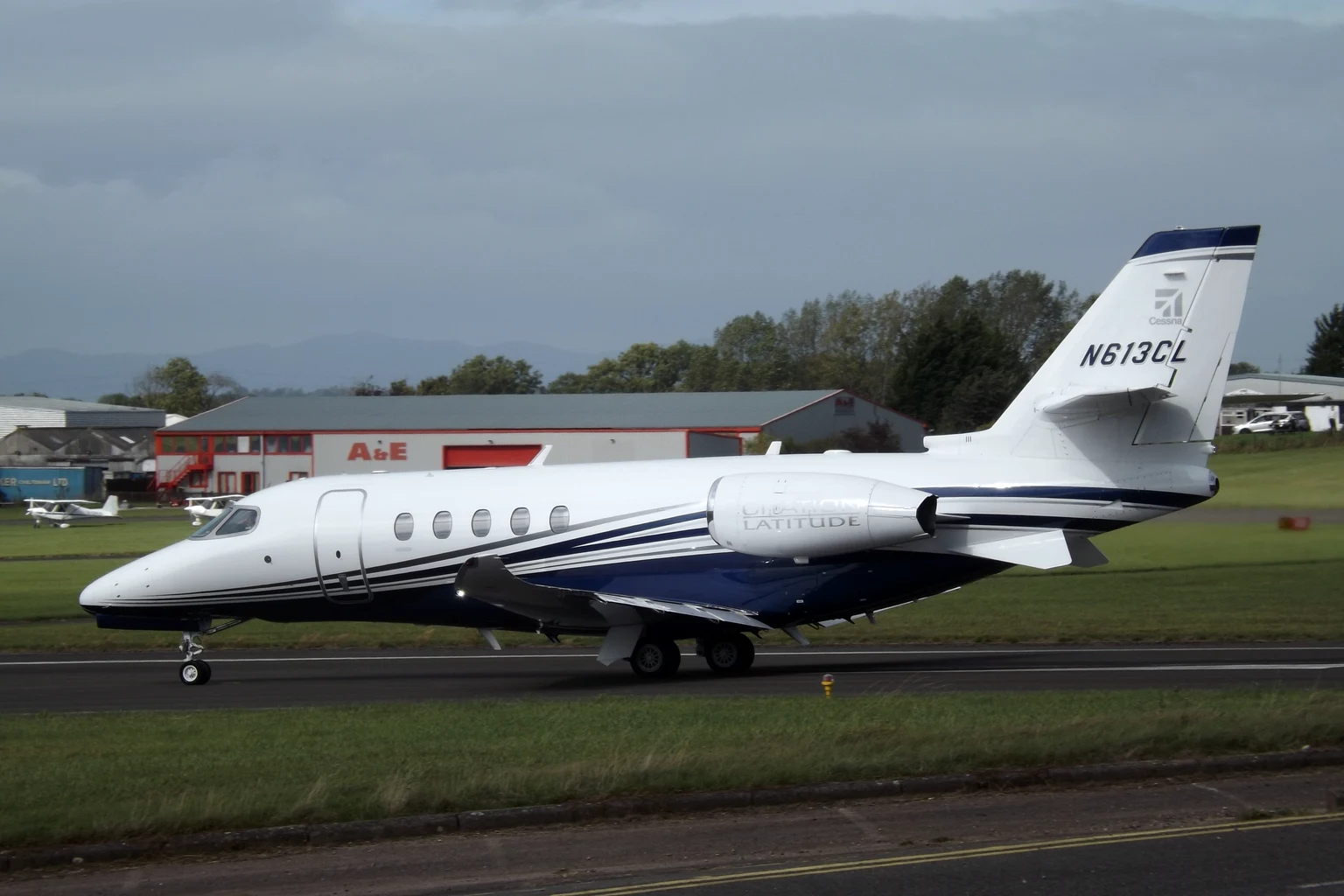 A white and navy Citation Latitude taxiing side-on at Gloucestershire Airport, hangars behind.