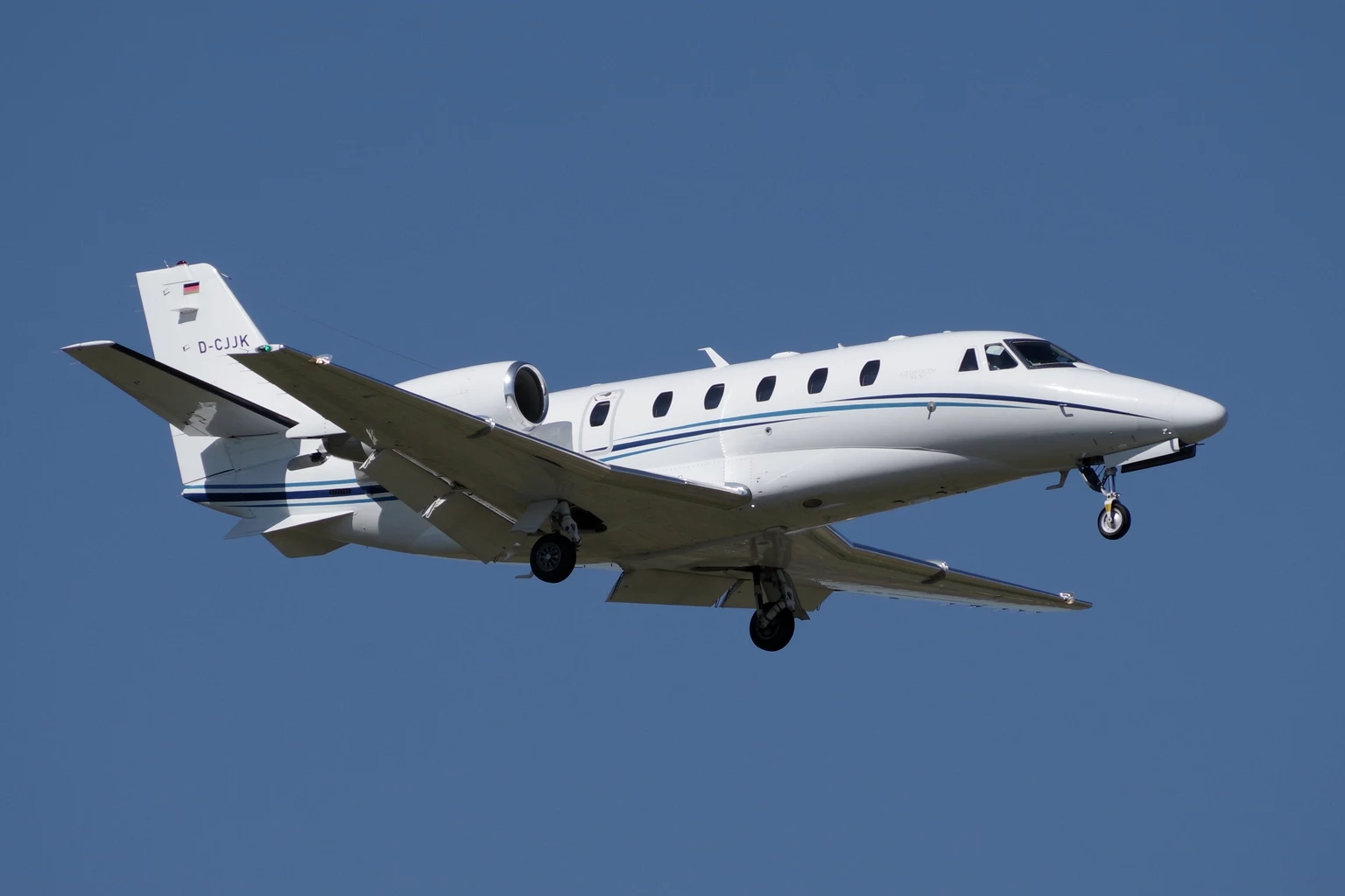 A white Cessna Citation XLS+ with a blue pinstripe climbing nose-high against a deep blue sky, seen from below at three-quarter.