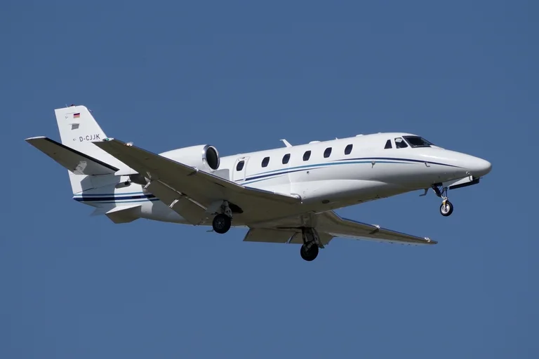 A white Cessna Citation XLS+ with a blue pinstripe climbing nose-high against a deep blue sky, seen from below at three-quarter.