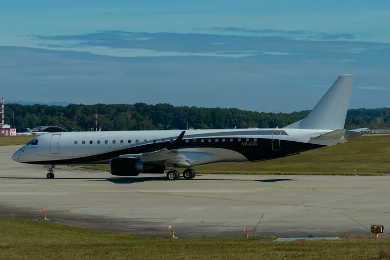 A white and black Embraer Lineage 1000 taxiing side-on at Geneva in low sun, trees and a blue sky behind.