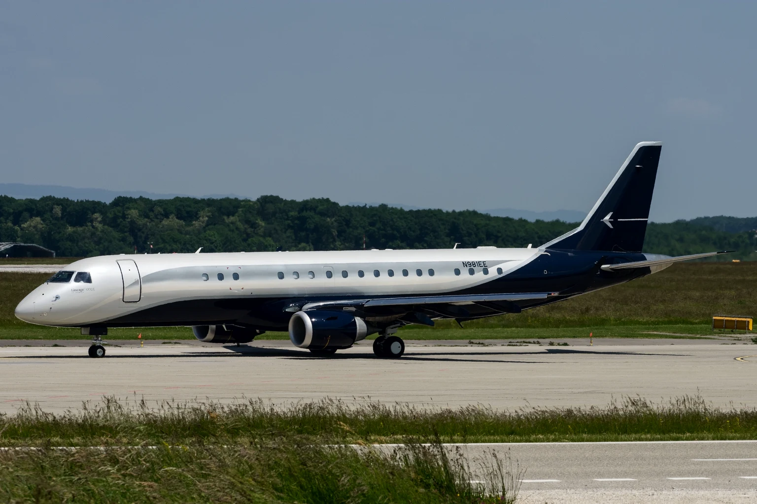 The Embraer-owned Lineage 1000 in silver with a navy tail and belly, taxiing side-on at Geneva.