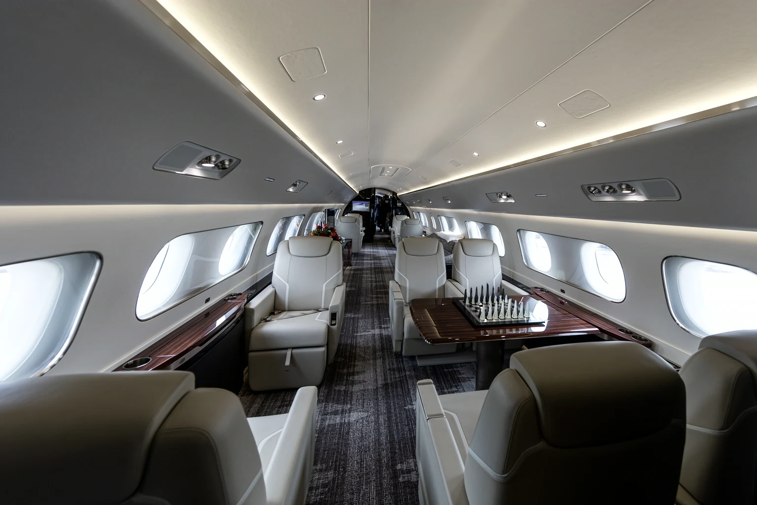 Looking down a Lineage 1000 cabin past cream leather club seats and a chess table on a dark wood console, long rows of windows.