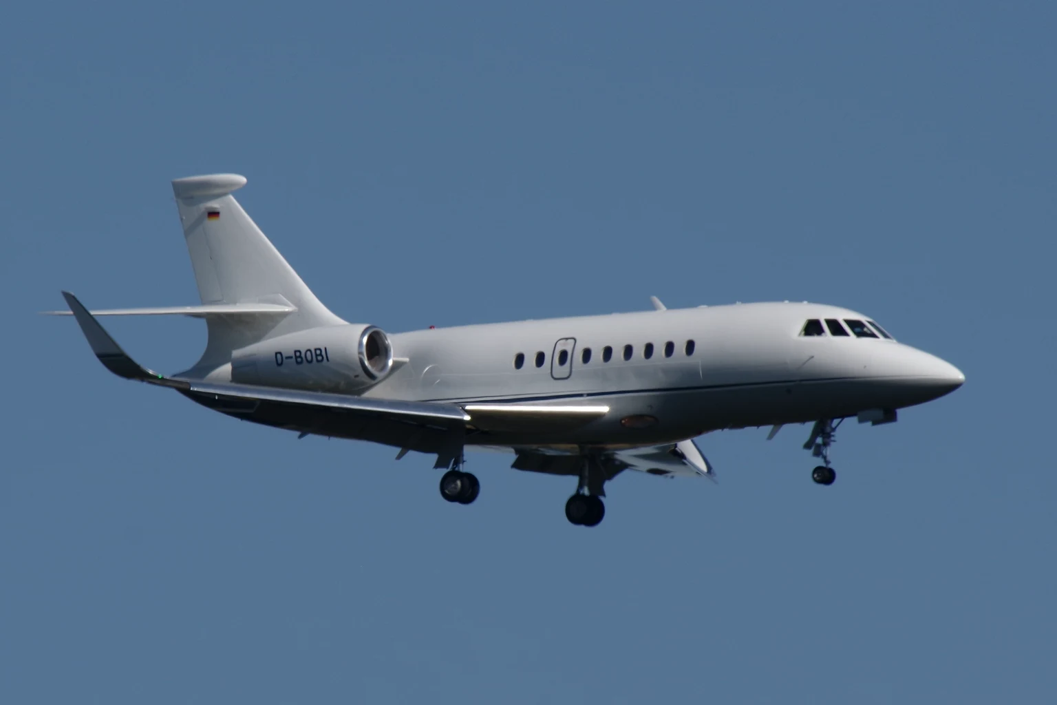 A plain white Dassault Falcon 2000LX with blended winglets on approach against a clear blue sky, three-quarter from the front.