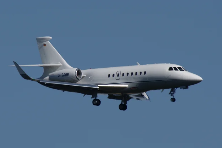 A plain white Dassault Falcon 2000LX with blended winglets on approach against a clear blue sky, three-quarter from the front.