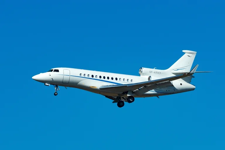 A white Dassault Falcon 7X with a thin blue swoosh on approach against a cloudless deep blue sky, side-on from slightly below.