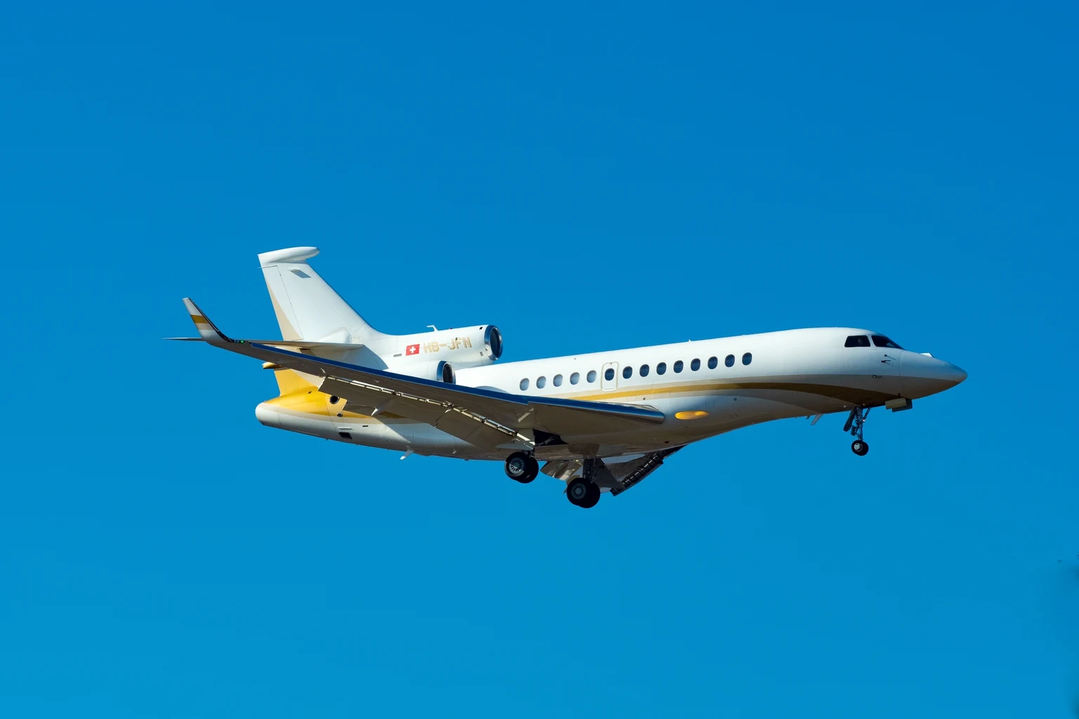 A white Falcon 7X with a gold belly band on approach against a deep blue sky.