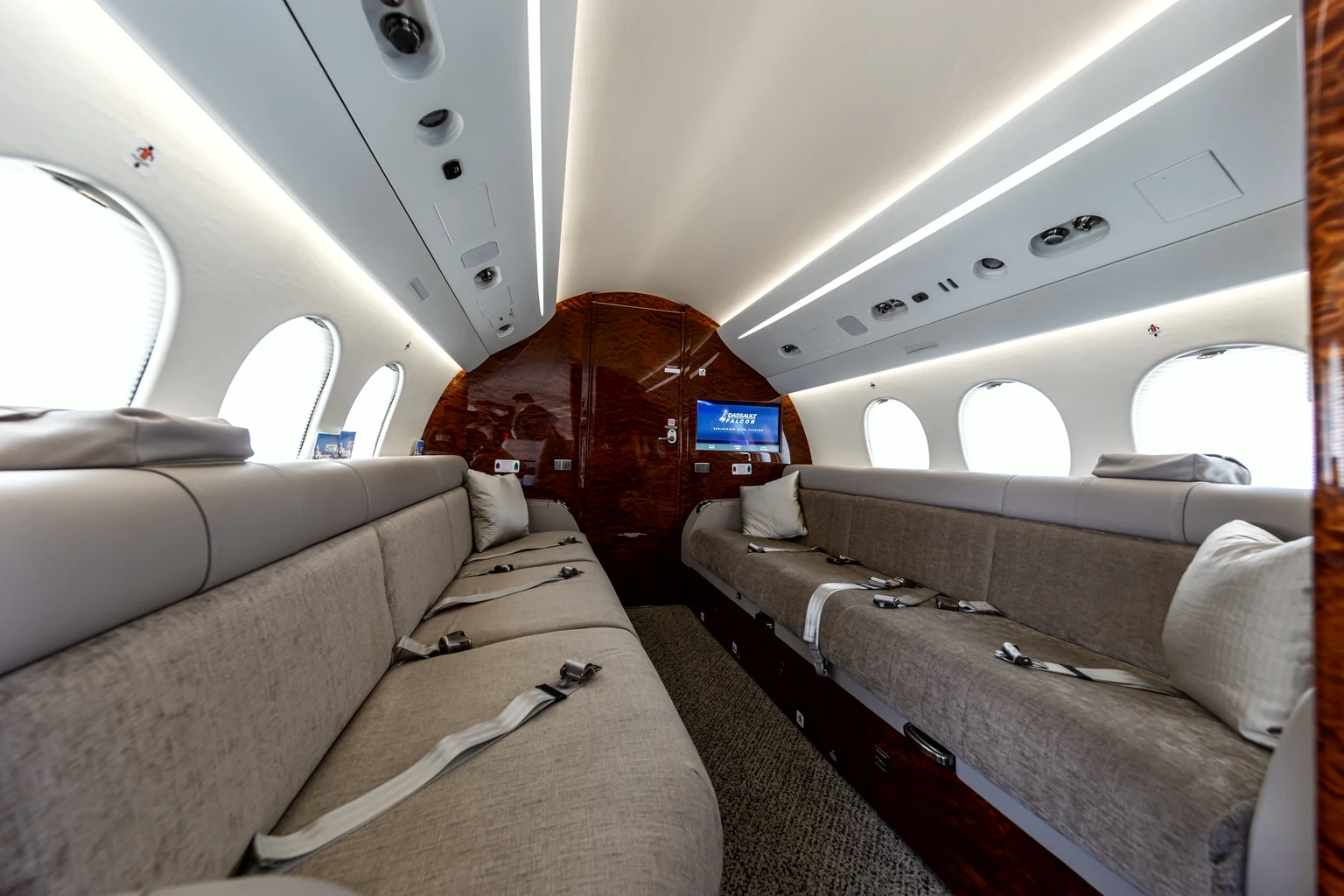 Twin beige divans facing each other down a Falcon 7X cabin toward a gloss burl-wood bulkhead, bright window light.
