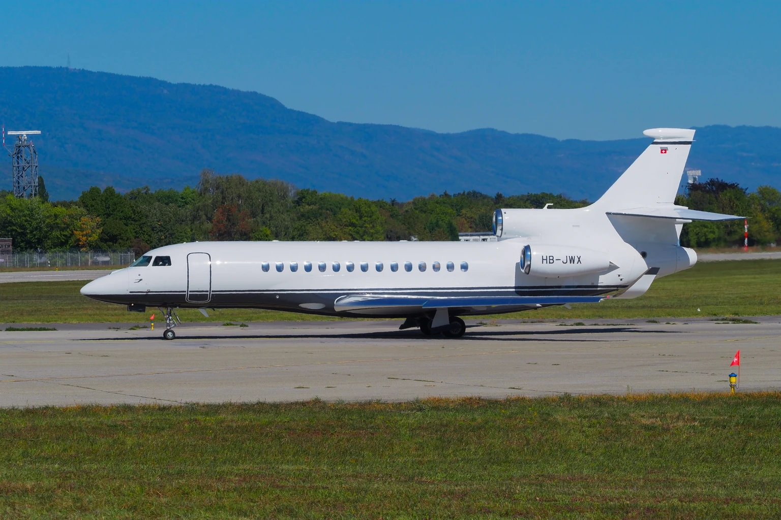 A white Dassault Falcon 8X with a navy cheatline taxiing side-on at Geneva in sunshine, the blue Jura ridge behind.