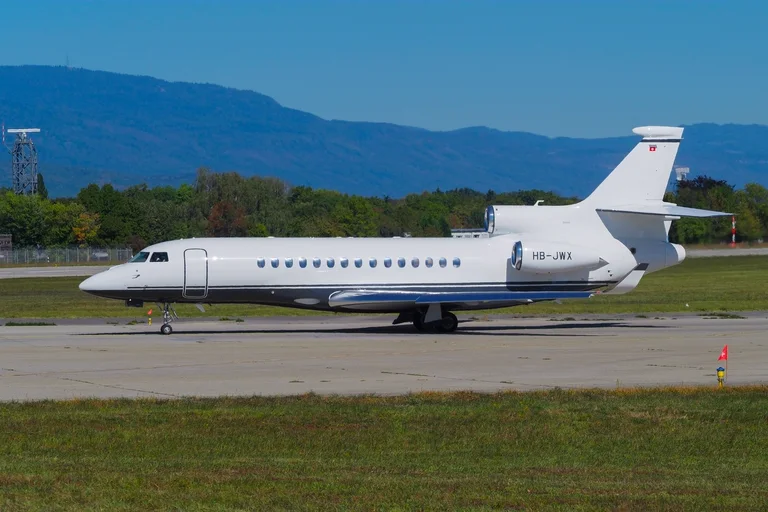 A white Dassault Falcon 8X with a navy cheatline taxiing side-on at Geneva in sunshine, the blue Jura ridge behind.