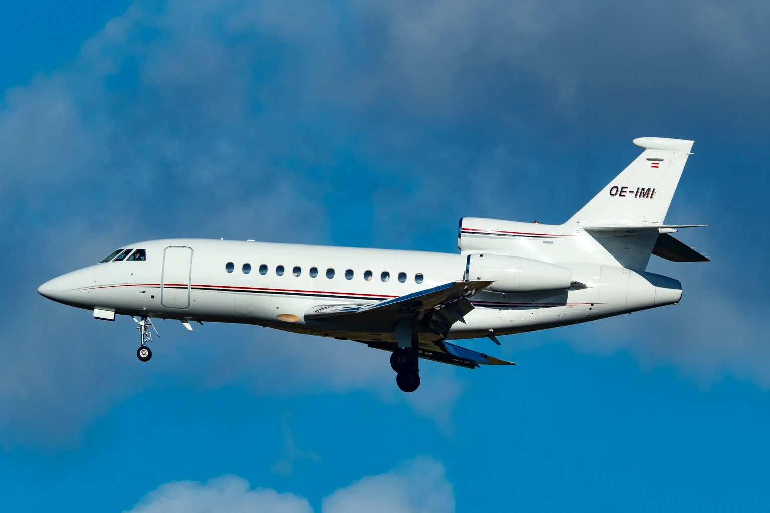 A white Dassault Falcon 900EX with red and navy pinstripes on approach at Geneva, side-on against a blue sky with broken cloud.