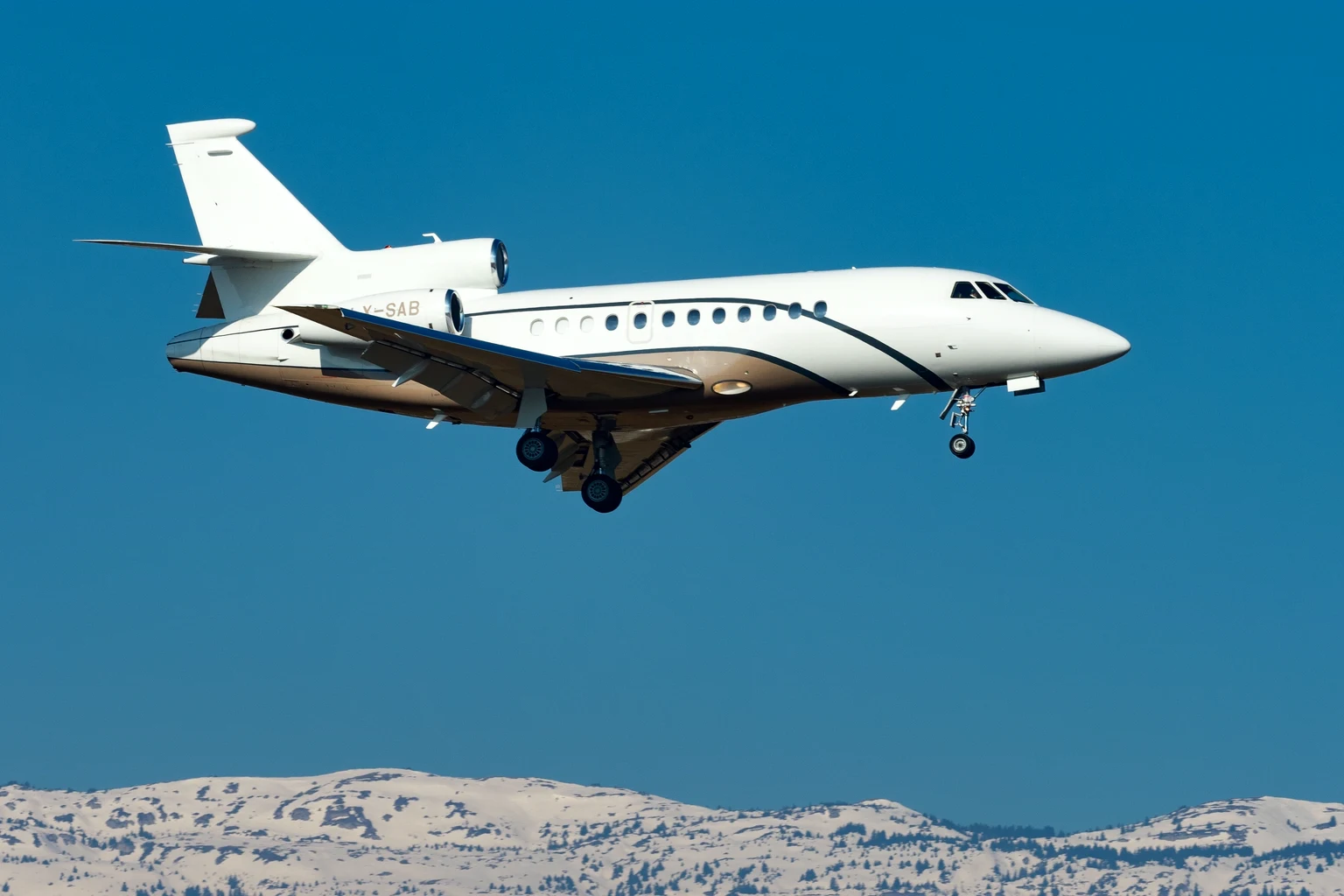 A white Falcon 900DX with a bronze belly and a dark cheatline on approach at Geneva, deep blue sky above the snow-covered Jura.
