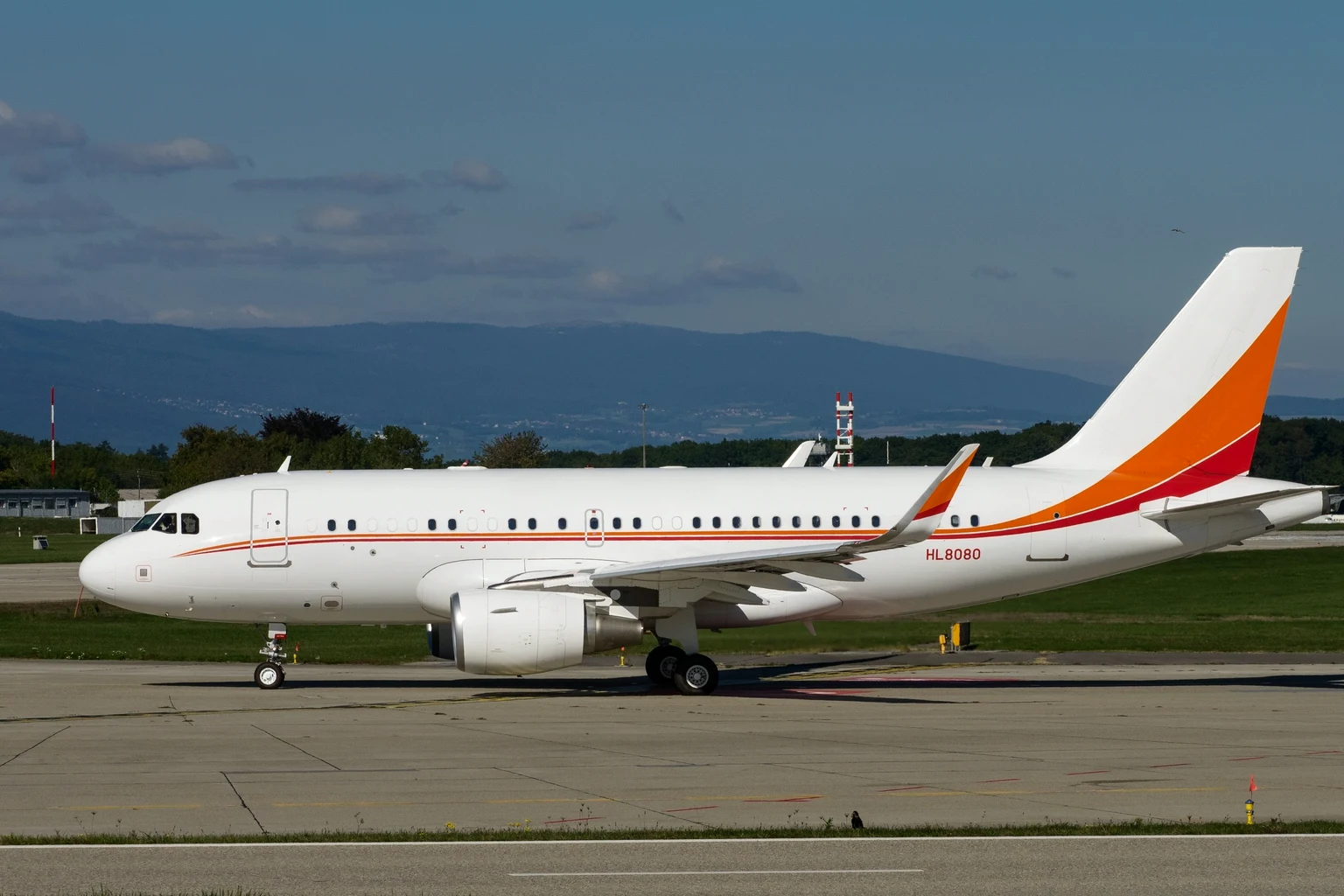 An Airbus ACJ319 corporate jet with sharklets, white with an orange and red tail, taxiing side-on at Geneva with the Jura on the horizon.