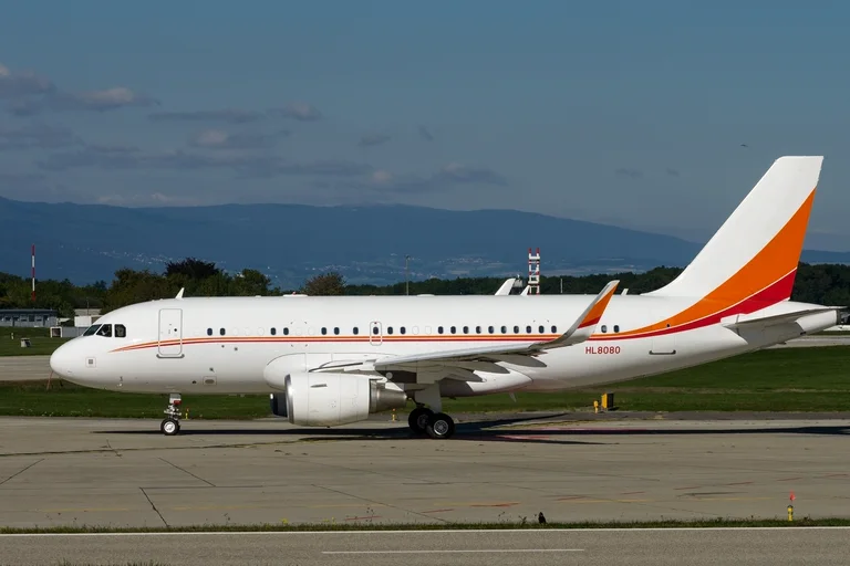 An Airbus ACJ319 corporate jet with sharklets, white with an orange and red tail, taxiing side-on at Geneva with the Jura on the horizon.