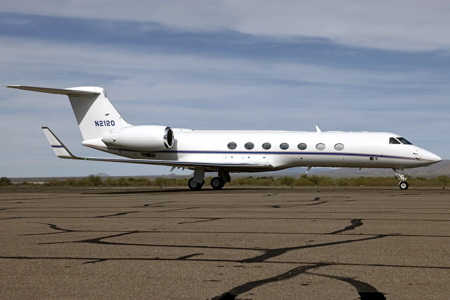 A white Gulfstream G550 with a blue and red cheatline taxiing side-on at Marana, Arizona, under a pale blue sky with desert hills behind.
