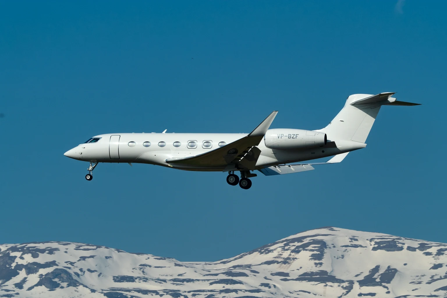 A plain white Gulfstream G650 on approach at Geneva against a deep blue sky, side-on, with snow-covered Alpine peaks along the bottom of the frame.