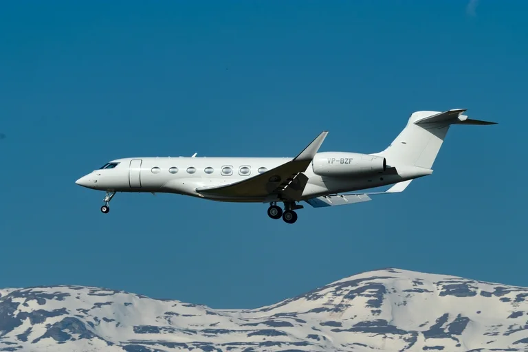 A plain white Gulfstream G650 on approach at Geneva against a deep blue sky, side-on, with snow-covered Alpine peaks along the bottom of the frame.
