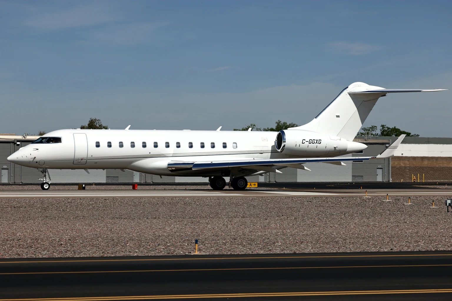 A plain white Bombardier Global 6000 with a thin grey cheatline on the runway at Scottsdale, side-on, under a blue sky with hangars behind.