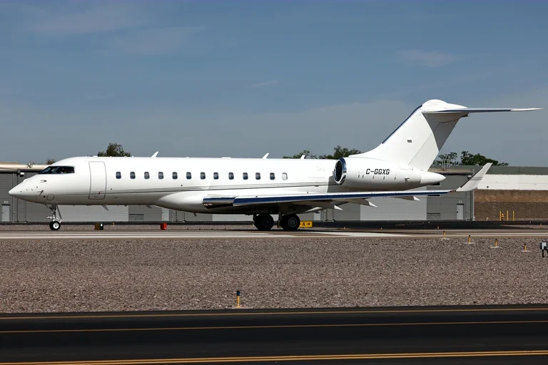 A plain white Bombardier Global 6000 with a thin grey cheatline on the runway at Scottsdale, side-on, under a blue sky with hangars behind.