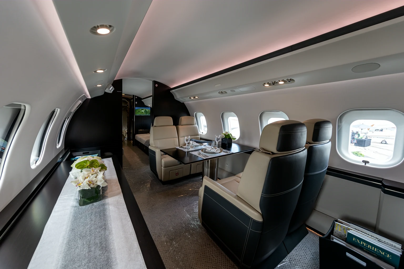 The middle of a Global 6000 cabin: a dining table laid for four between cream and black leather seats, a credenza with white orchids, and a divan at the far end.