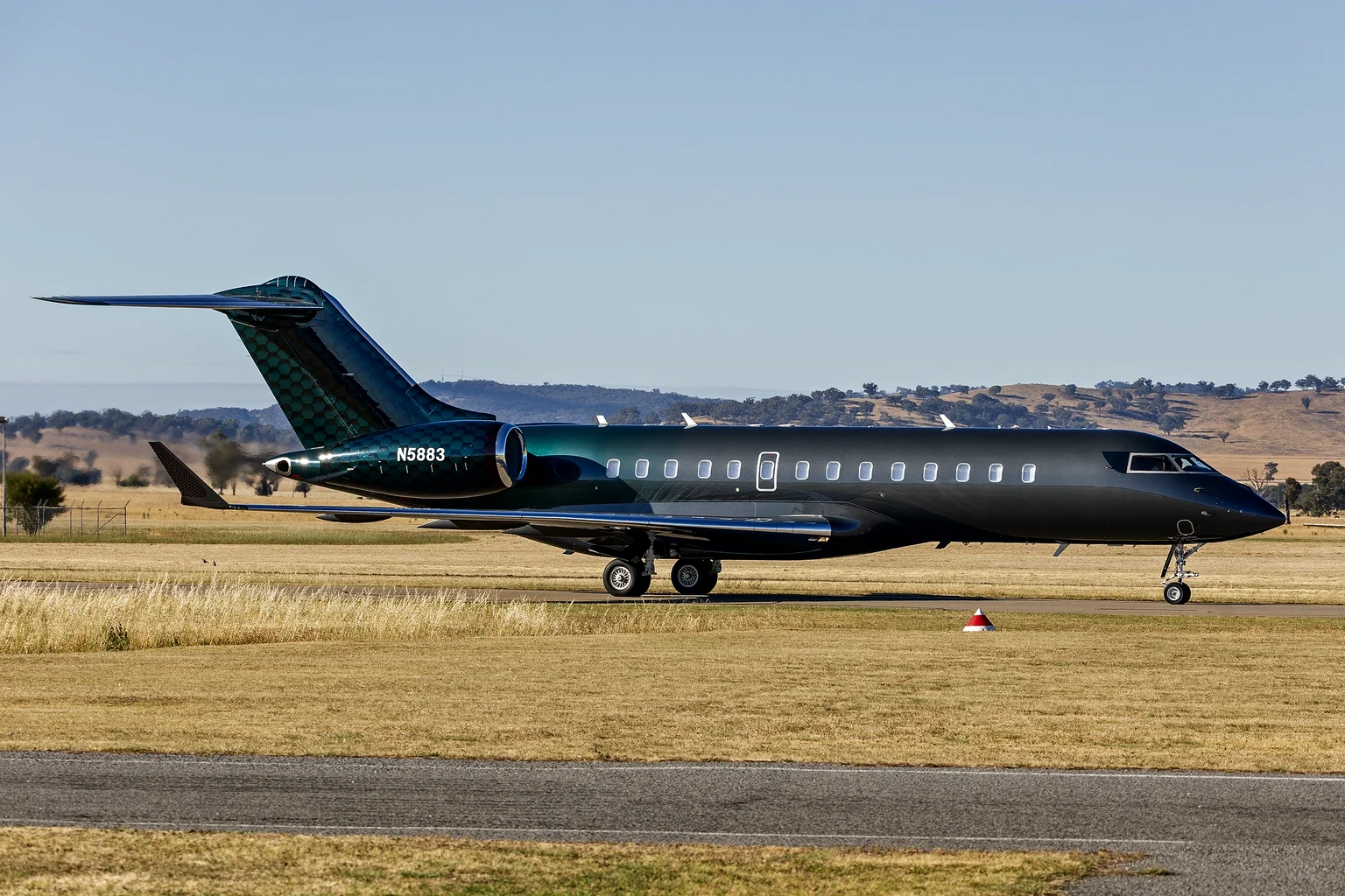 A black Bombardier Global 6500 with a teal scale-pattern tail taxiing side-on in bright sun at Wagga Wagga, dry grass and hills behind.