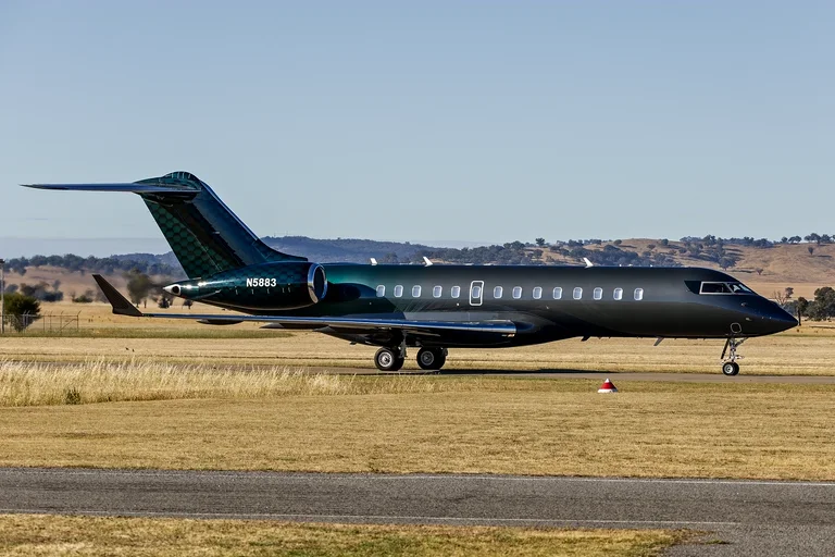 A black Bombardier Global 6500 with a teal scale-pattern tail taxiing side-on in bright sun at Wagga Wagga, dry grass and hills behind.