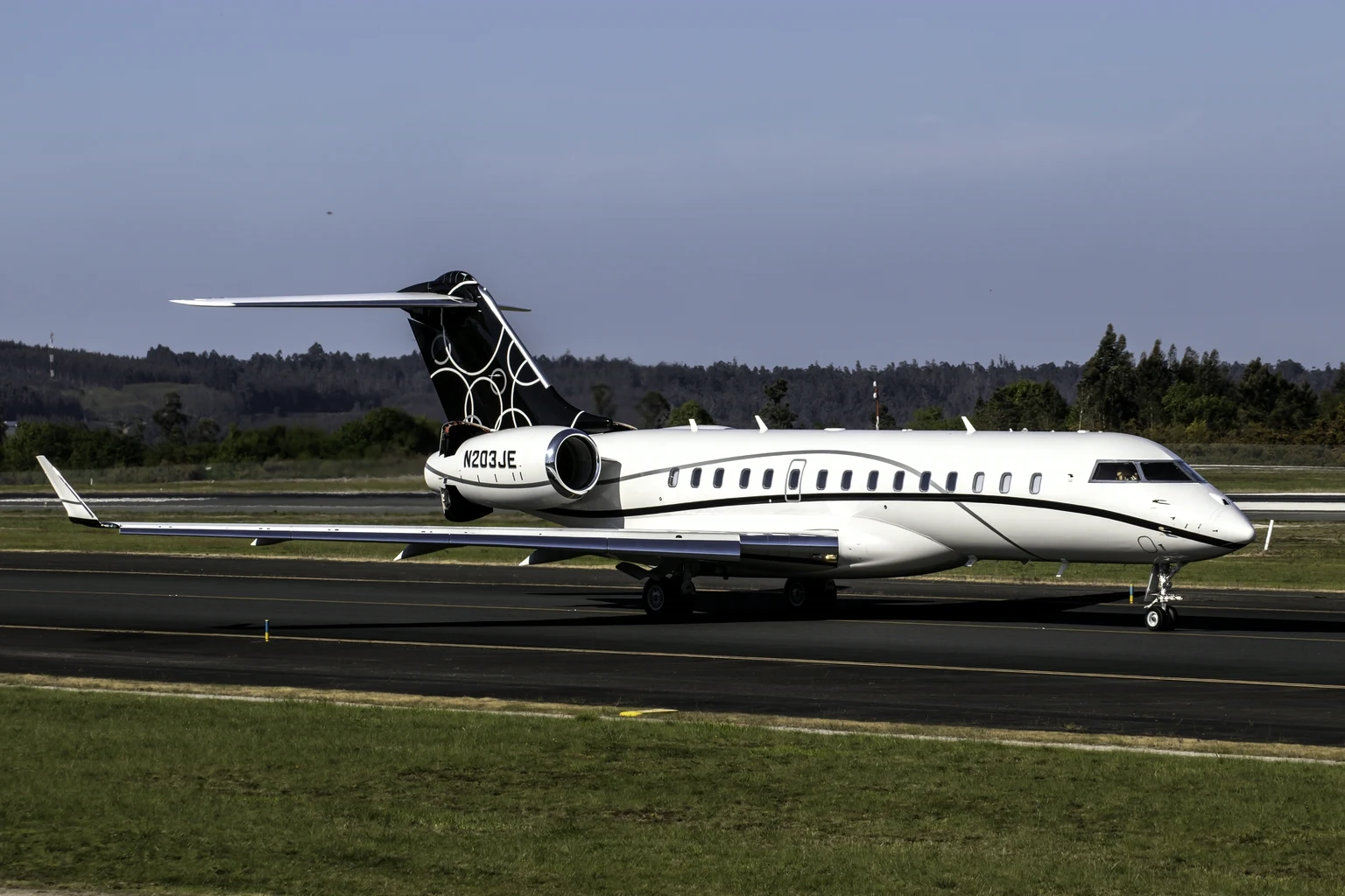 A privately owned Bombardier Global 7500, white with a black tail patterned in white rings, taxiing side-on at Santiago de Compostela under a dusky sky, pine woods behind.