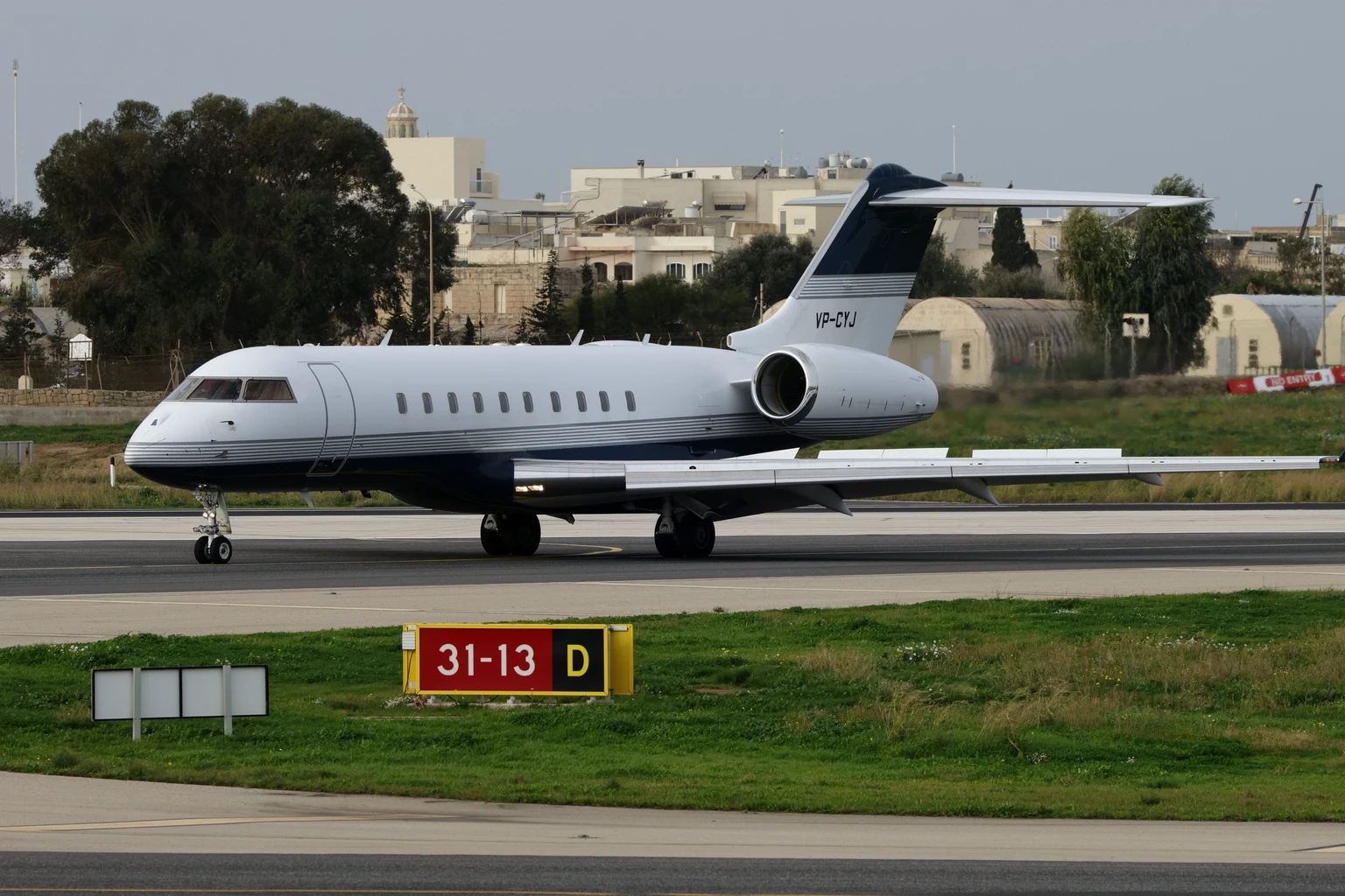 A Bombardier Global business jet with a dark blue belly taxiing past hangars