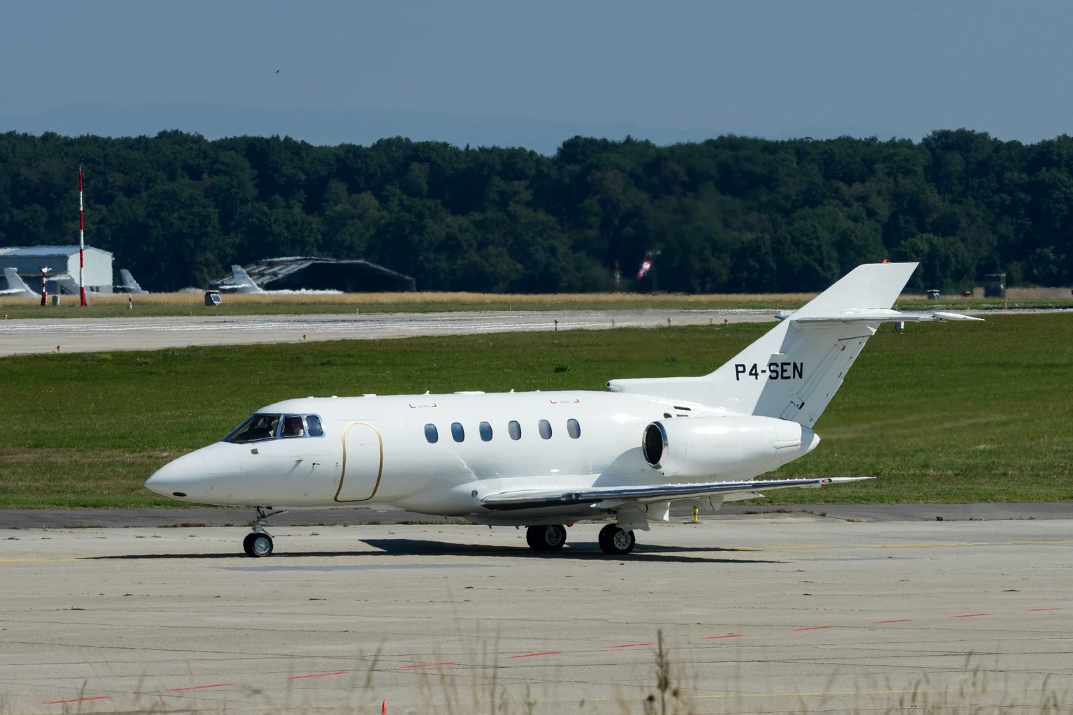 A plain white Hawker 800XP with a gold line around its door taxiing side-on at Geneva in summer sun, woods behind the airfield.